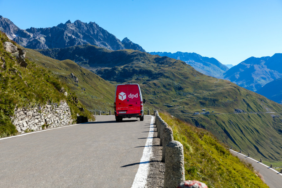 	Czerwony samochód dostawczy DPD jedzie wzdłuż górskiej drogi z widokiem na majestatyczne Alpy.